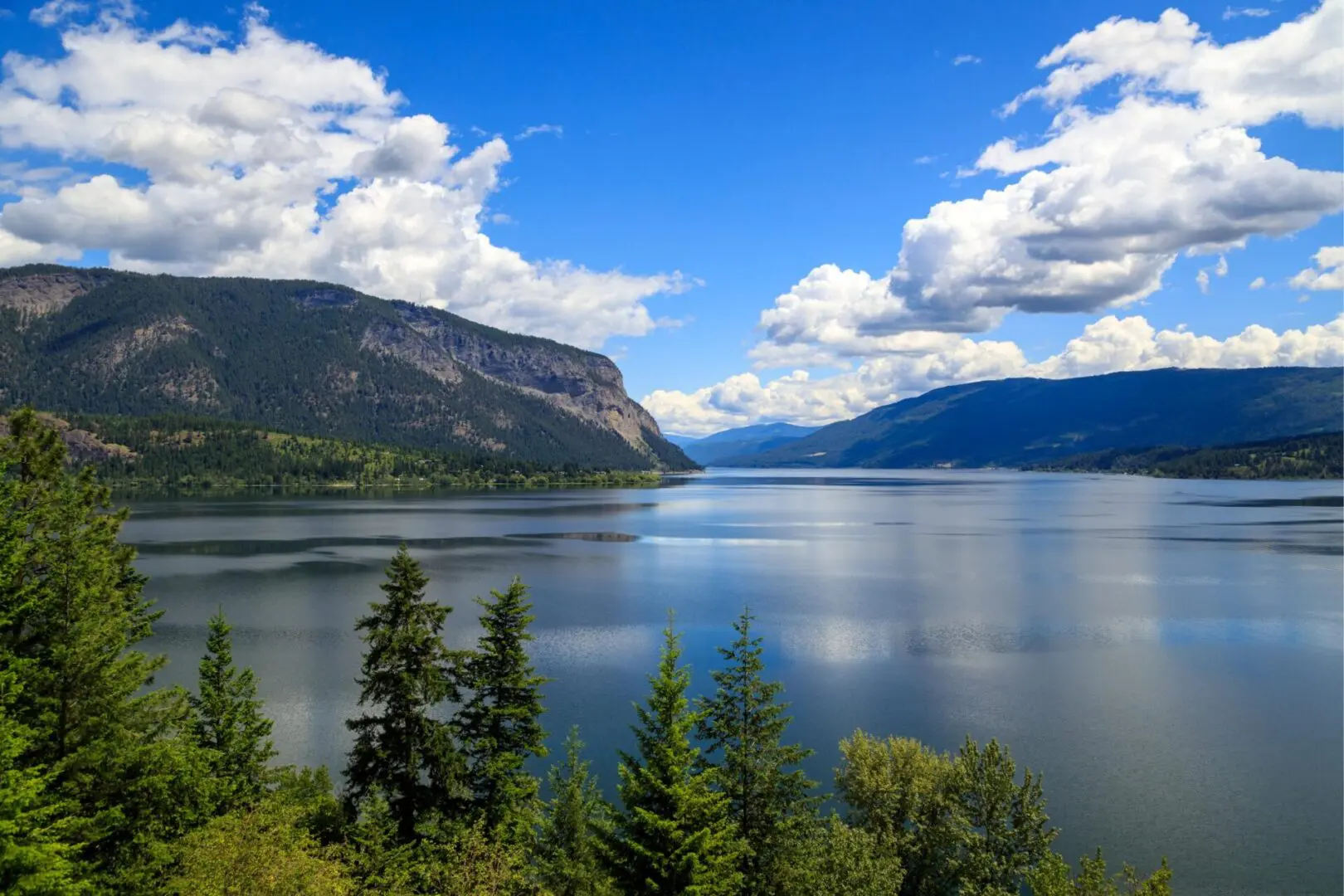 A serene lake surrounded by lush green forests and distant mountains under a bright blue sky with scattered clouds.
